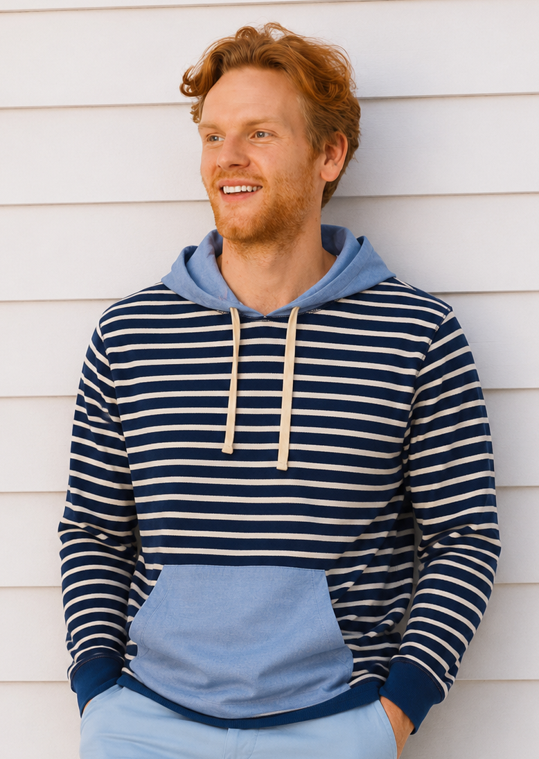 A smiling man with red hair and a beard leans against a white horizontal siding wall, wearing a color-blocked hoodie with navy and white horizontal stripes on the top half, a light blue color-blocked bottom half, and a light blue hood. He is also wearing light blue shorts.