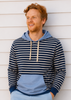 A smiling man with red hair and a beard leans against a white horizontal siding wall, wearing a color-blocked hoodie with navy and white horizontal stripes on the top half, a light blue color-blocked bottom half, and a light blue hood. He is also wearing light blue shorts.