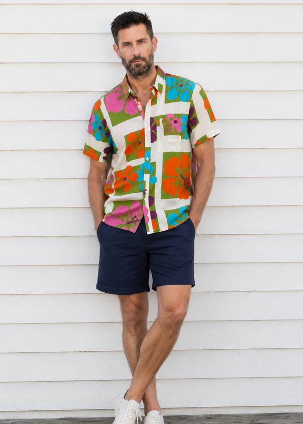 A man wearing a colorful short-sleeve button-up shirt with a patchwork tropical print, navy blue shorts, and white sneakers, standing against a white wooden wall.