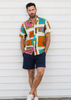 A man wearing a colorful short-sleeve button-up shirt with a patchwork tropical print, navy blue shorts, and white sneakers, standing against a white wooden wall.