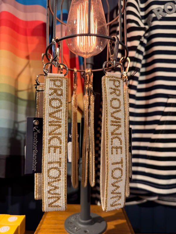 A close-up of beaded keychains with 'PROVINCETOWN' text hanging from a display stand in a retail setting. The keychains feature cream and tan beading with gold-toned lettering, alongside a black tag from AuditorFashion.