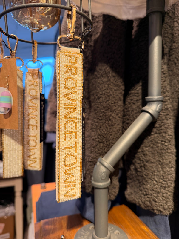 A close-up of a woven rectangular keychain or bag charm with the words "PROVINCETOWN" in raised tan lettering on a cream fabric background, hanging from a metal display stand in a retail setting.