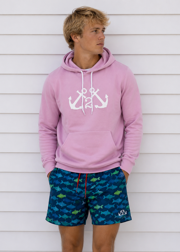 Person wearing a pink hoodie with anchor design and blue swim shorts with fish pattern against a white wall.