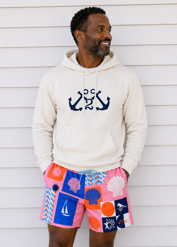 Man wearing a white hoodie with anchor design and colorful shorts with sea-themed patterns against a wooden wall.
