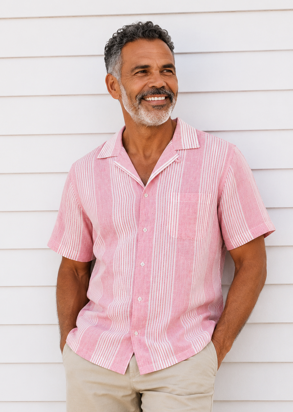 A smiling man with gray hair and a beard wearing a pink and white vertically striped short-sleeve camp collar shirt and beige shorts, standing against a white horizontal siding wall.