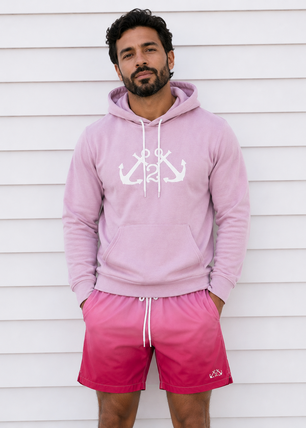 Man wearing a pink hoodie and shorts with a white anchor design against a white wooden background