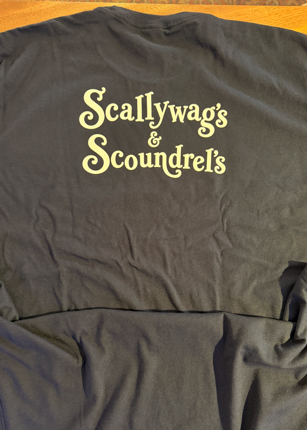 A dark charcoal gray t-shirt laid flat, featuring the words 'Scallywag's & Scoundrel's' printed in a light yellow gothic font across the chest.