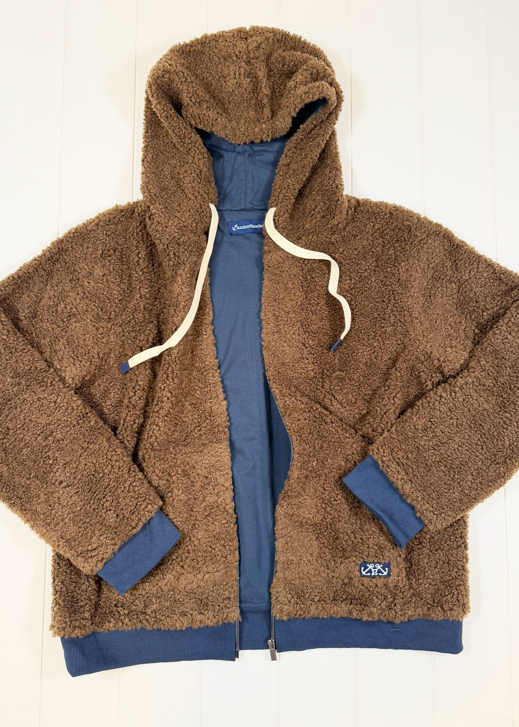 A brown sherpa fleece zip-up hoodie jacket with a blue inner lining, cream-colored drawstrings, and a small white logo patch on the lower left pocket, laid flat on a white wood background.