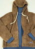 A brown sherpa fleece zip-up hoodie jacket with cream drawstrings, layered over a blue zippered inner jacket. The hood is up, and the jacket is laid flat against a white wood-paneled background.