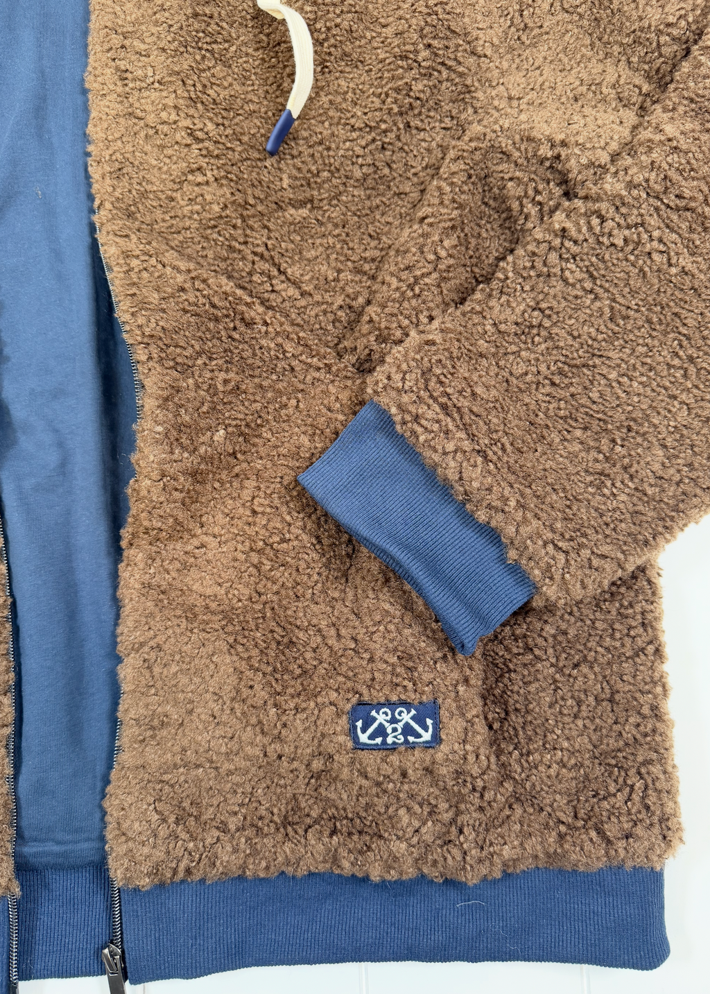Close-up of a brown sherpa fleece zip-up jacket with blue ribbed trim, cuffs, and hem, featuring a small embroidered anchor logo patch on the lower front.