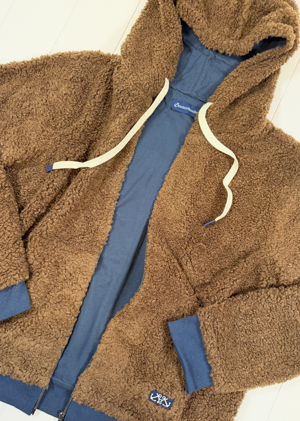 A brown teddy fleece zip-up hoodie jacket layered over a blue zip-up sweatshirt. The outer fleece has cream-colored drawstrings, and a small embroidered logo patch is visible near the lower left pocket.