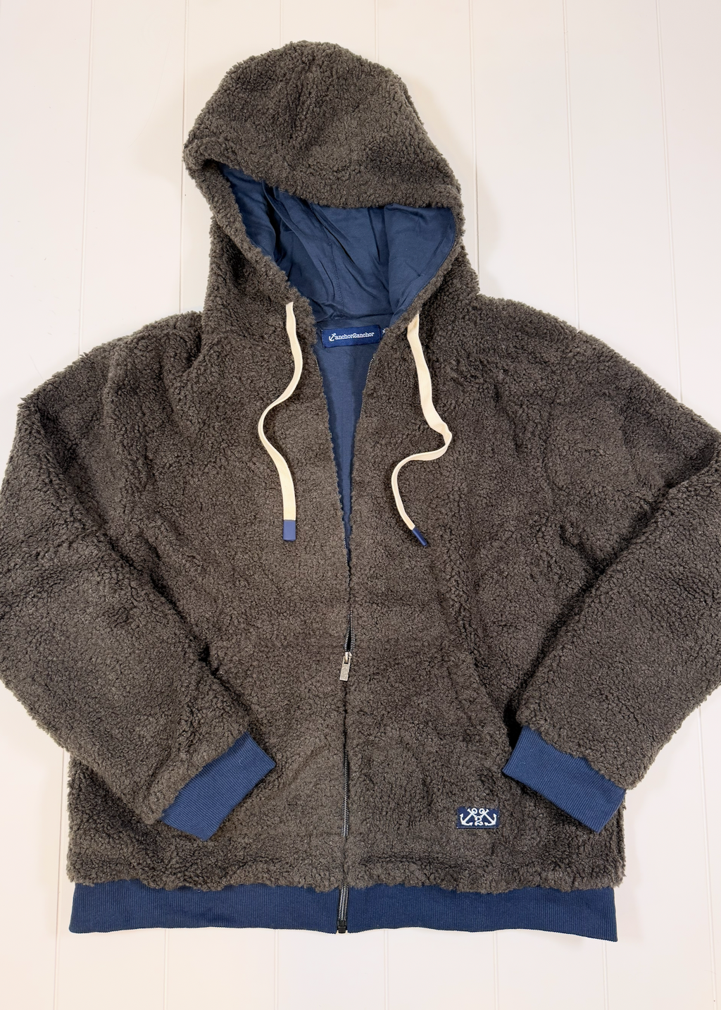 A dark brown sherpa fleece zip-up hoodie jacket with a navy blue lining, cream-colored drawstrings, and navy blue ribbed cuffs and hem, laid flat on a white wood panel background.