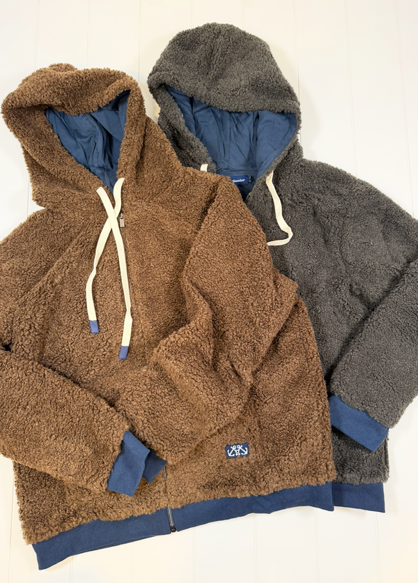 Two teddy fleece zip-up hoodies laid flat on a white wood background, one in brown and one in dark gray, both with blue lining and white drawstrings.