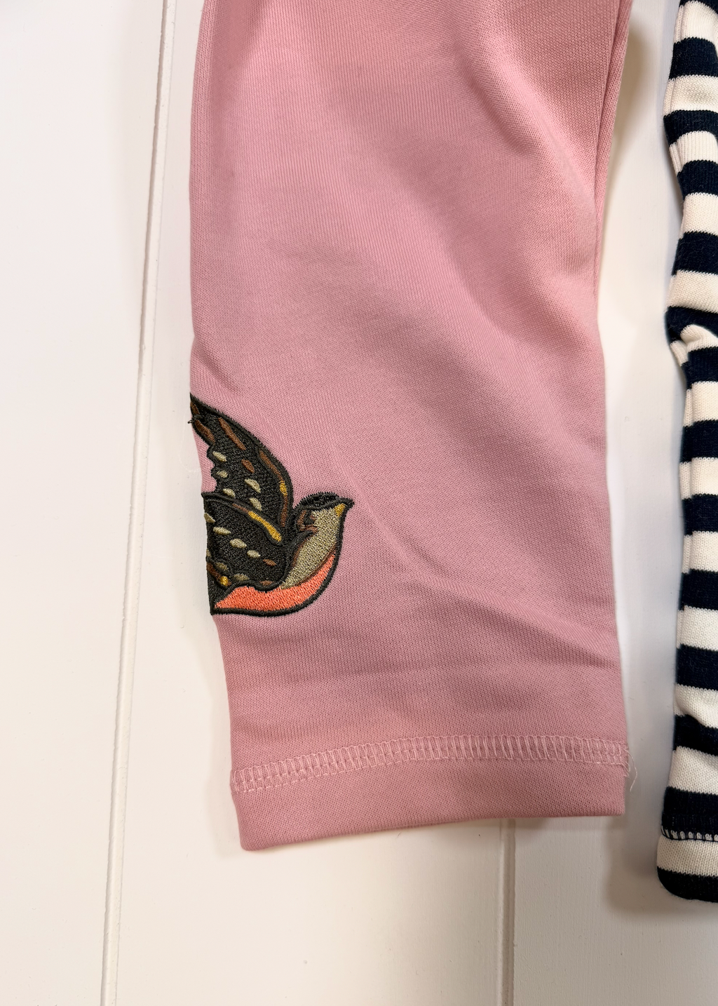 A close-up of a pink garment, likely a baby tights or pants leg, featuring an embroidered bird motif near the lower cuff. A portion of a navy and white striped garment is visible on the right edge.