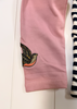 A close-up of a pink garment, likely a baby tights or pants leg, featuring an embroidered bird motif near the lower cuff. A portion of a navy and white striped garment is visible on the right edge.