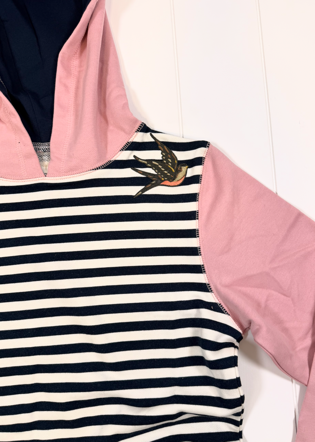 A close-up of a hooded top with a navy and white striped body, pink hood and sleeve, and a small embroidered swallow motif near the collar.