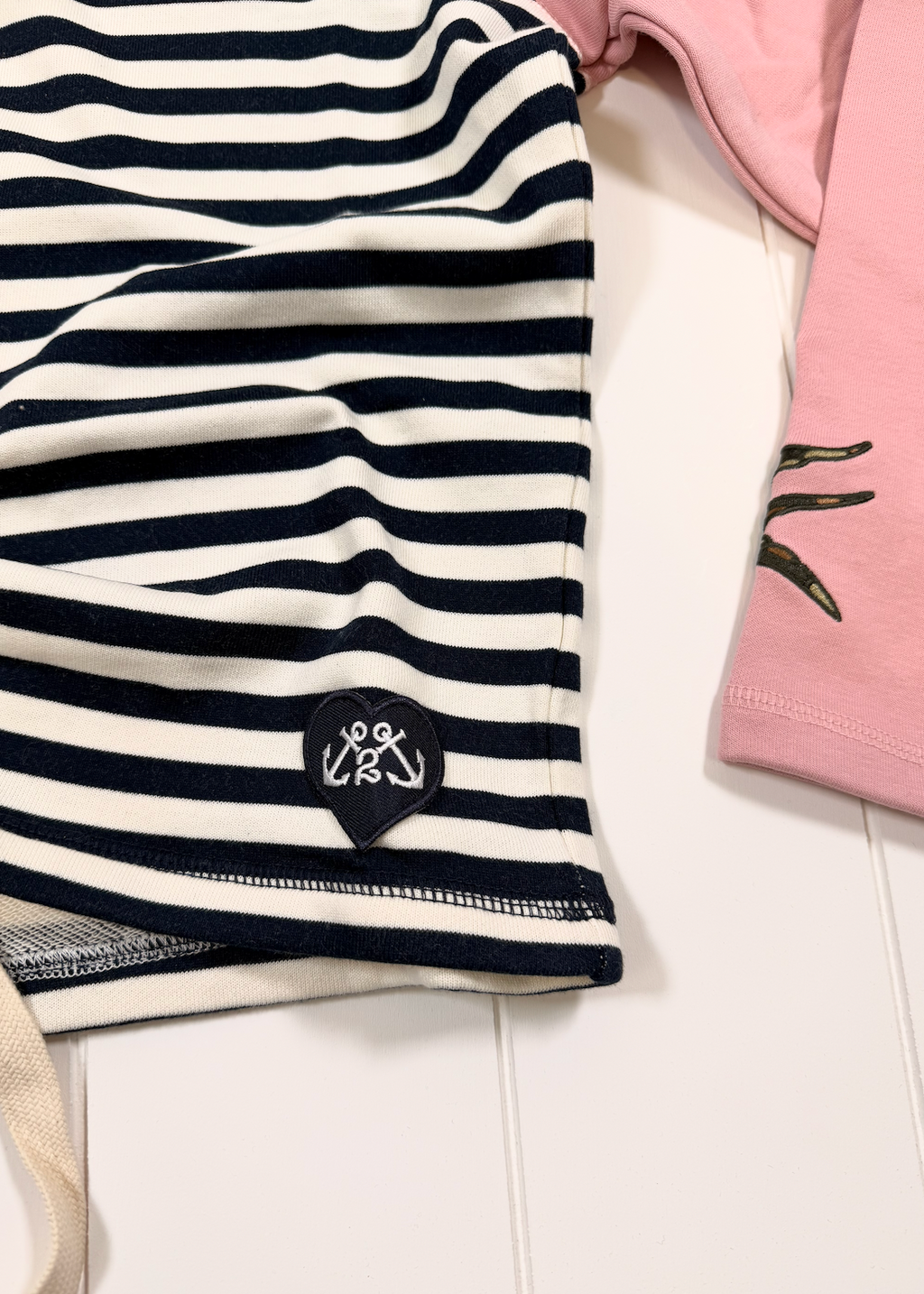 A close-up of a navy and white horizontally striped garment laid flat on a white surface, with a small black embroidered shield logo featuring two white anchors. Part of a pink garment is visible at the right edge.