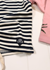 A close-up of a navy and white horizontally striped garment laid flat on a white surface, with a small black embroidered shield logo featuring two white anchors. Part of a pink garment is visible at the right edge.