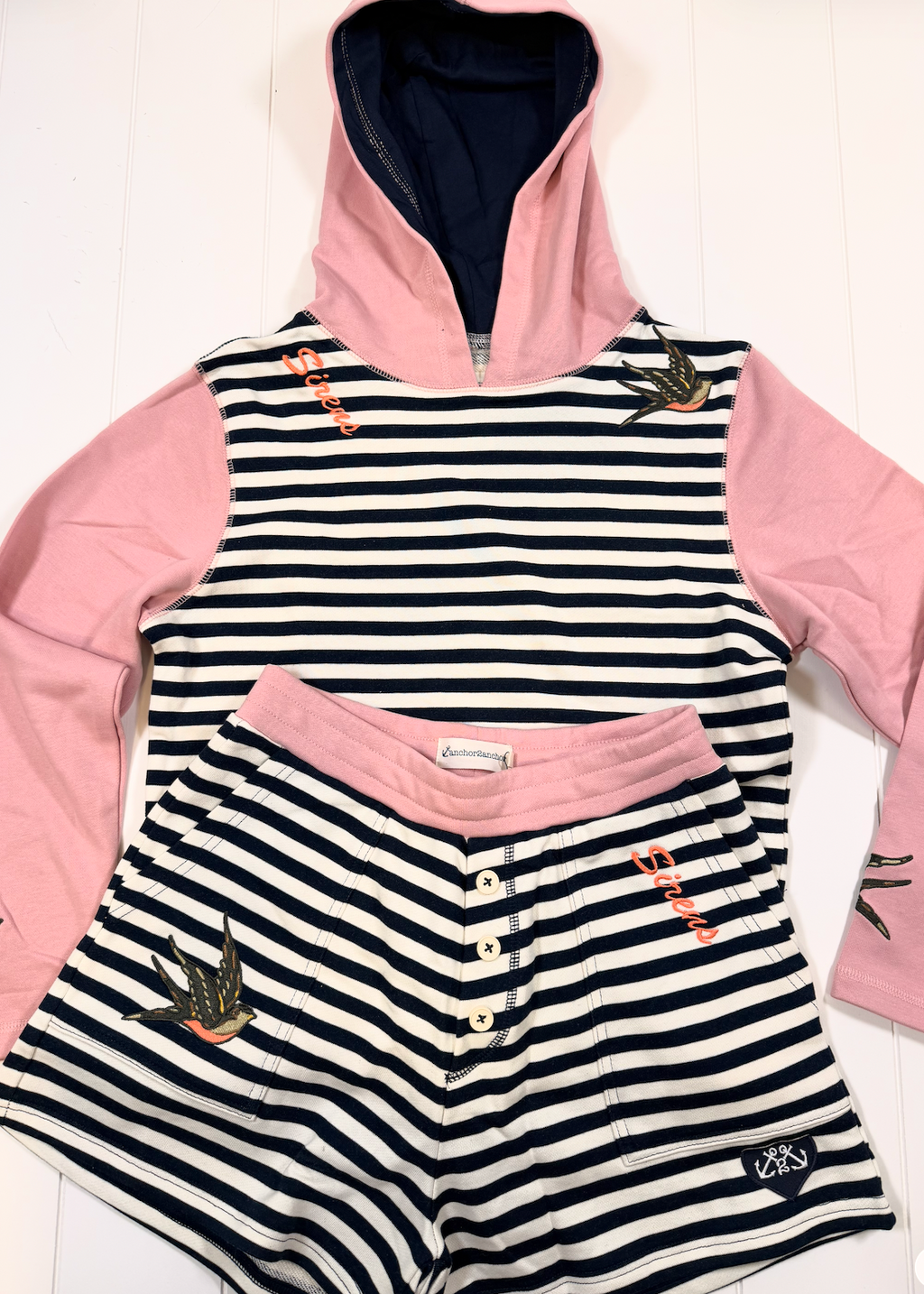 A children's two-piece loungewear set featuring a hooded long-sleeve top and matching shorts. The top has pink sleeves and hood lining, a navy and white horizontal stripe body, and embroidered swallow motifs.
