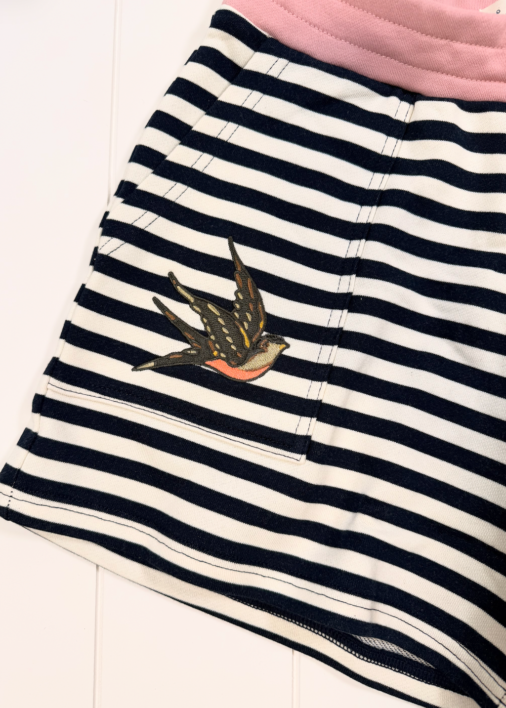 A close-up of a children's striped garment, likely a skirt or shorts, featuring horizontal navy and white stripes, a pink waistband, and a embroidered swallow motif on the front pocket.