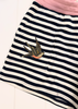 A close-up of a children's striped garment, likely a skirt or shorts, featuring horizontal navy and white stripes, a pink waistband, and a embroidered swallow motif on the front pocket.