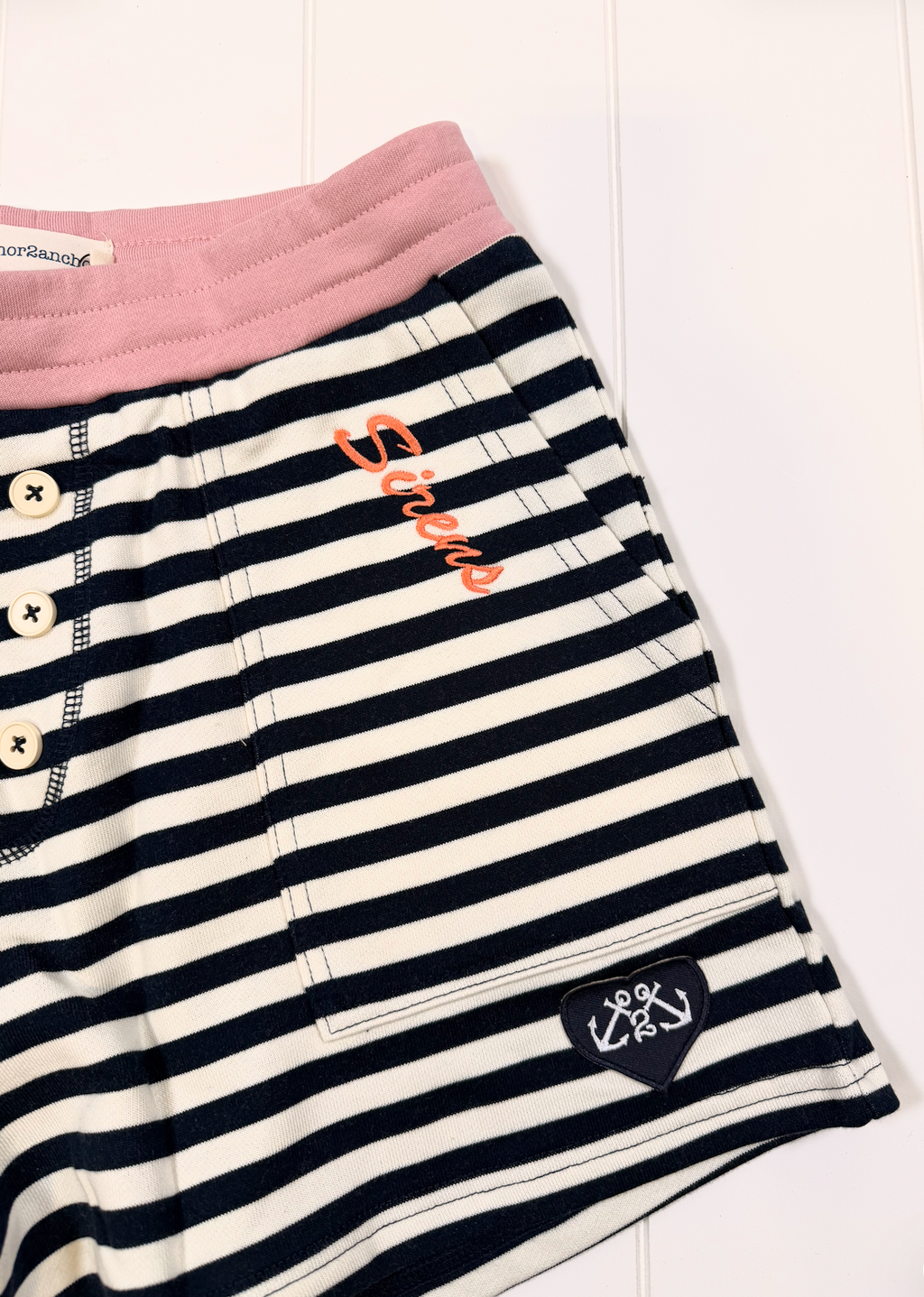 Close-up of striped shorts with a pink waistband, white buttons, orange script embroidery, and a small black heart patch on a white background.