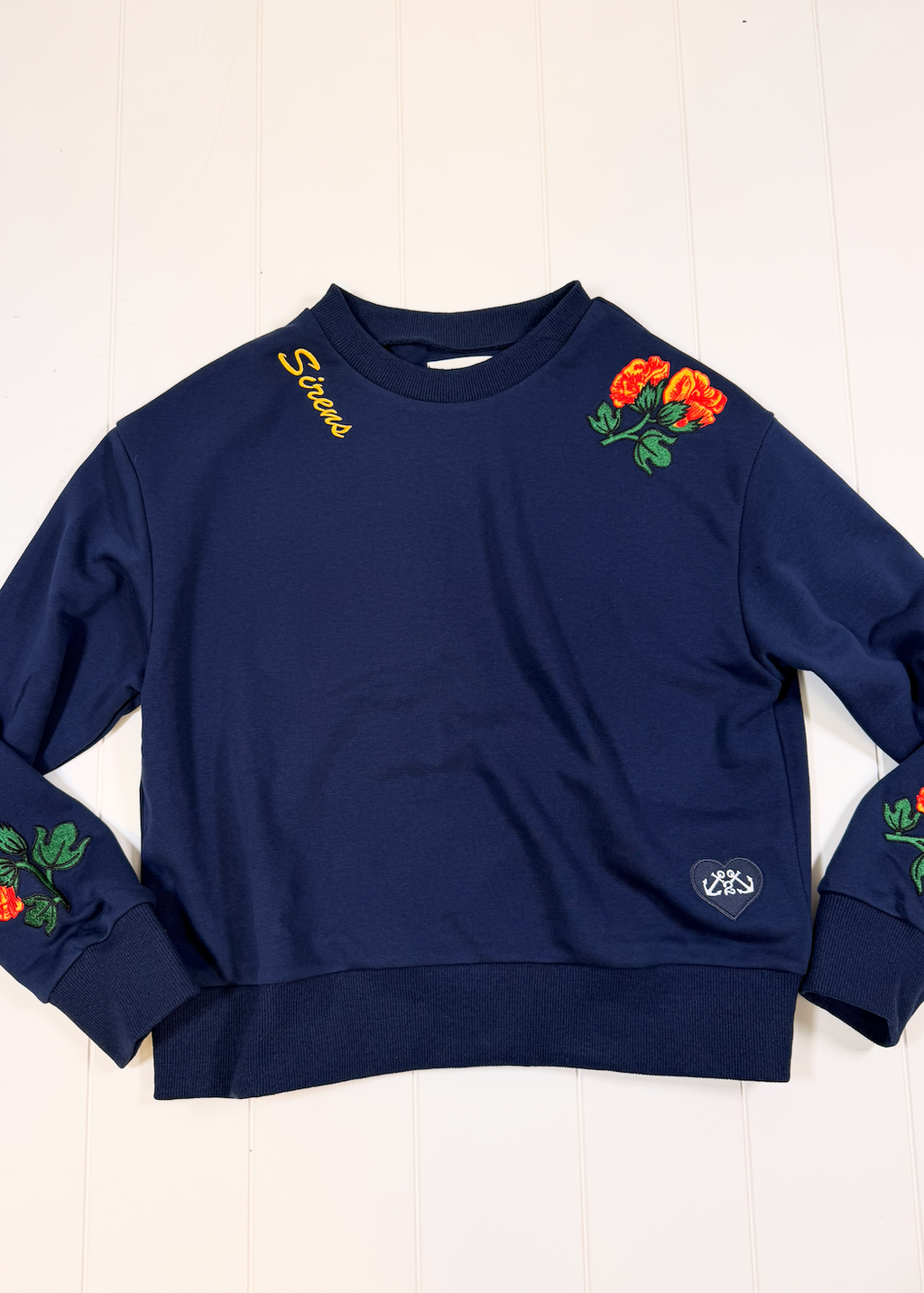 A navy blue crewneck sweatshirt with embroidered orange roses and green leaves on the shoulders and cuffs, featuring yellow 'Stans' embroidery on the left shoulder and a small circular logo patch on the right sleeve, laid flat on a white wood background.
