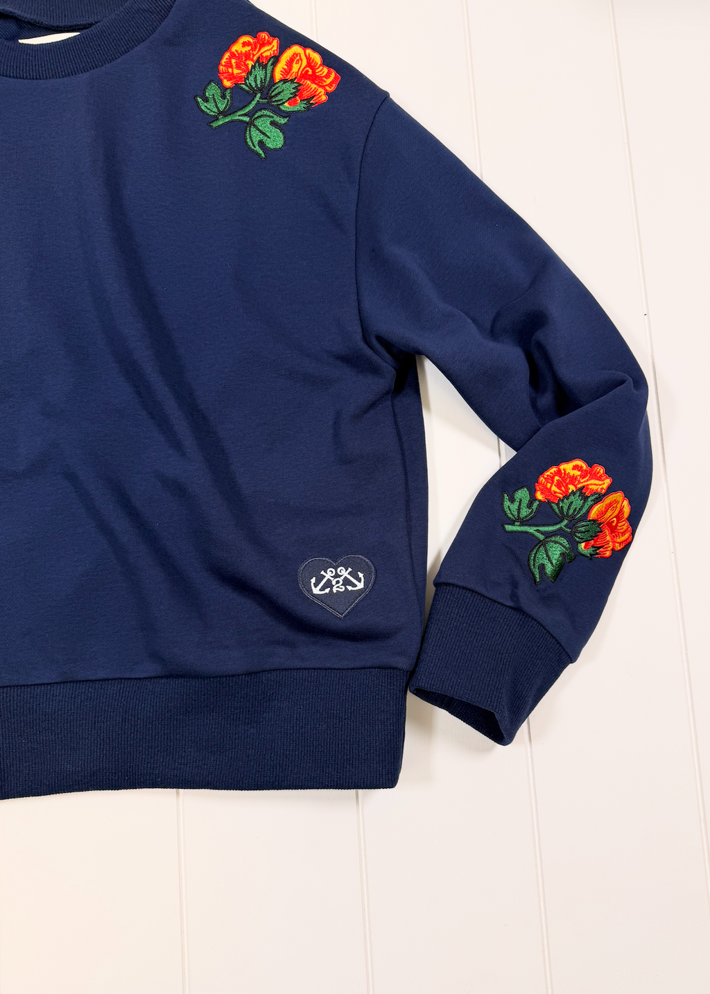 A navy blue crewneck sweatshirt featuring embroidered orange roses with green leaves on the left chest and left sleeve, plus a small heart-shaped embroidered patch on the lower left sleeve, laid flat on a white wood background.