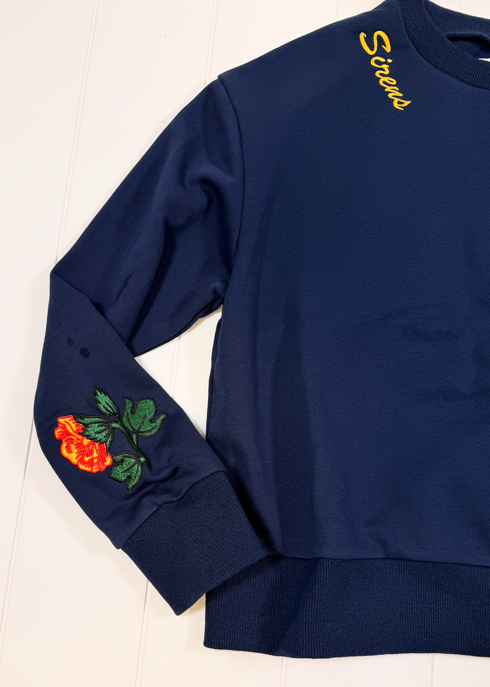A navy blue crewneck sweatshirt laid flat on a white wooden background, featuring a yellow embroidered 'Scouts' logo on the right shoulder and a colorful embroidered red rose with green leaves on the left sleeve.
