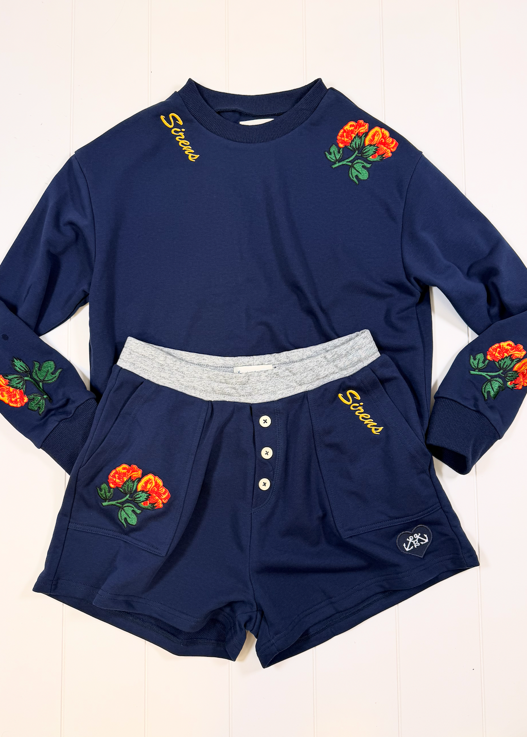 A navy blue two-piece loungewear set featuring a long-sleeve top and matching shorts. The top has yellow 'Stories' embroidery on the chest, orange rose floral patches on the shoulders and sleeves, and a contrasting light gray elastic waistband.
