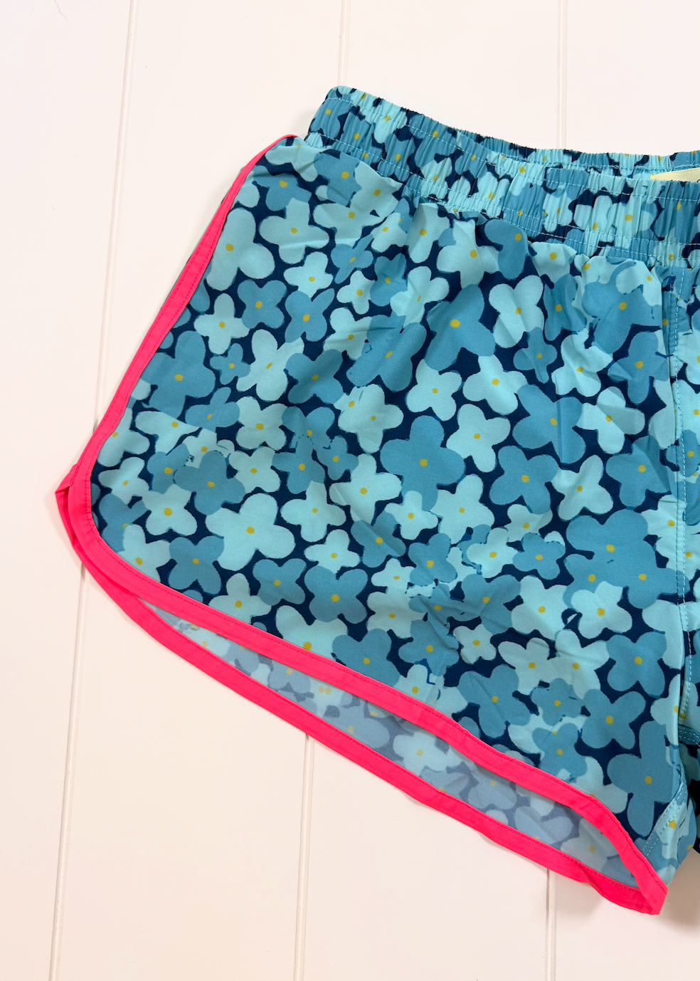 A pair of women's athletic shorts featuring a blue and light blue floral pattern, with a vibrant pink trim along the side and hem, laid flat on a white wood-paneled background.