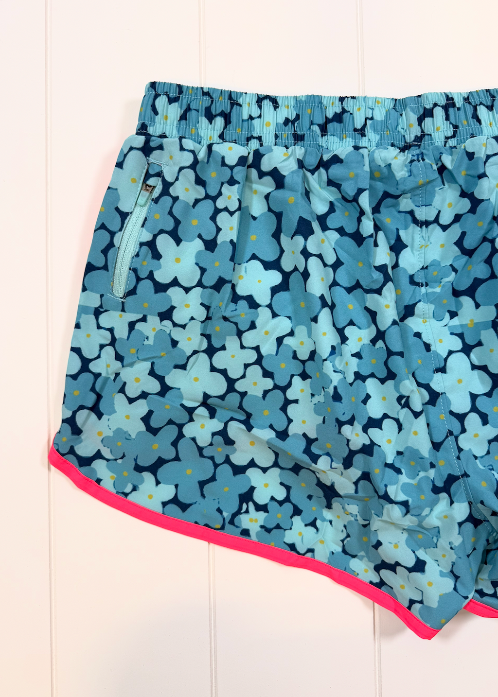 Blue floral print athletic shorts with a pink trim, laid flat on a white paneled background.