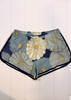 A pair of women's shorts featuring a large-scale floral print with navy blue, light blue, and cream flowers outlined in black, finished with a contrasting navy blue hem.