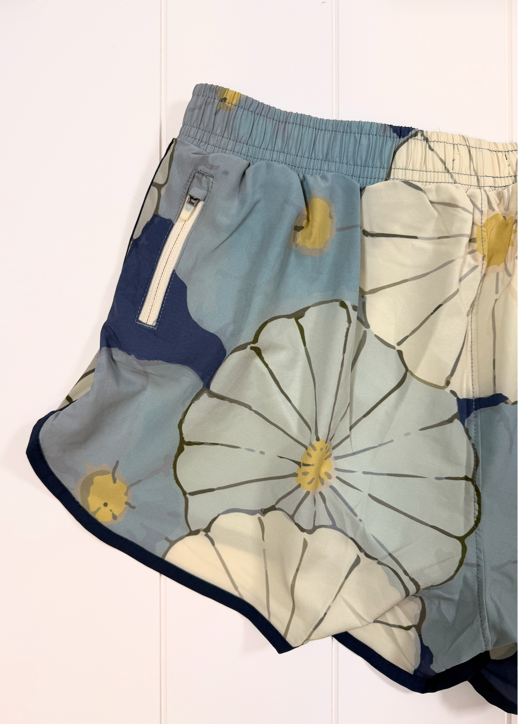 Light blue and navy floral print shorts with a white zipper pocket, laid flat against a white paneled background.