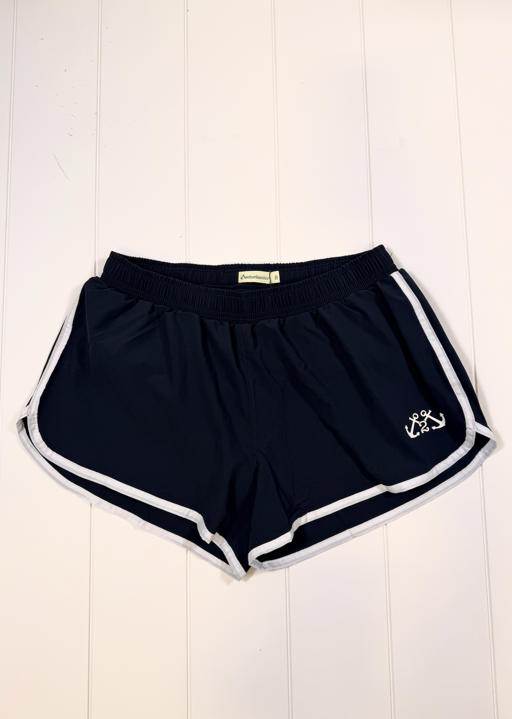 Navy blue athletic shorts with white side stripes and a white embroidered anchor logo on the left leg, laid flat on a white wood plank background.