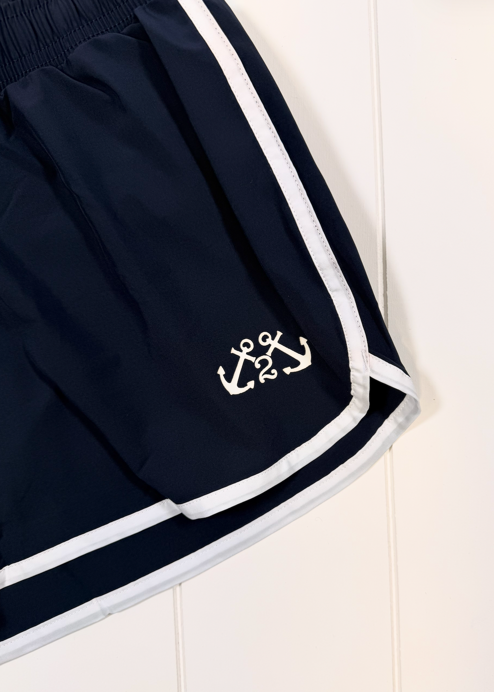 Close-up of navy blue athletic shorts with white side stripes and a white anchor logo near the hem, laid against a white paneled background.
