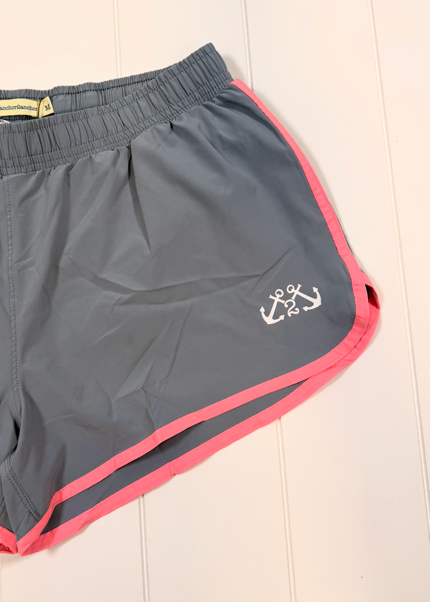 Gray athletic shorts with pink trim and a white anchor logo, laid flat on a light wood background.