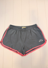 Gray athletic shorts with pink trim and a white logo on the left leg, laid flat on a white wood plank background.