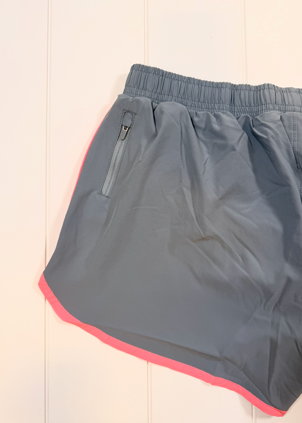 Gray athletic shorts with a bright pink inner hem and a zippered side pocket, laid flat on a white paneled background.