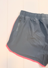 Gray athletic shorts with a bright pink inner hem and a zippered side pocket, laid flat on a white paneled background.