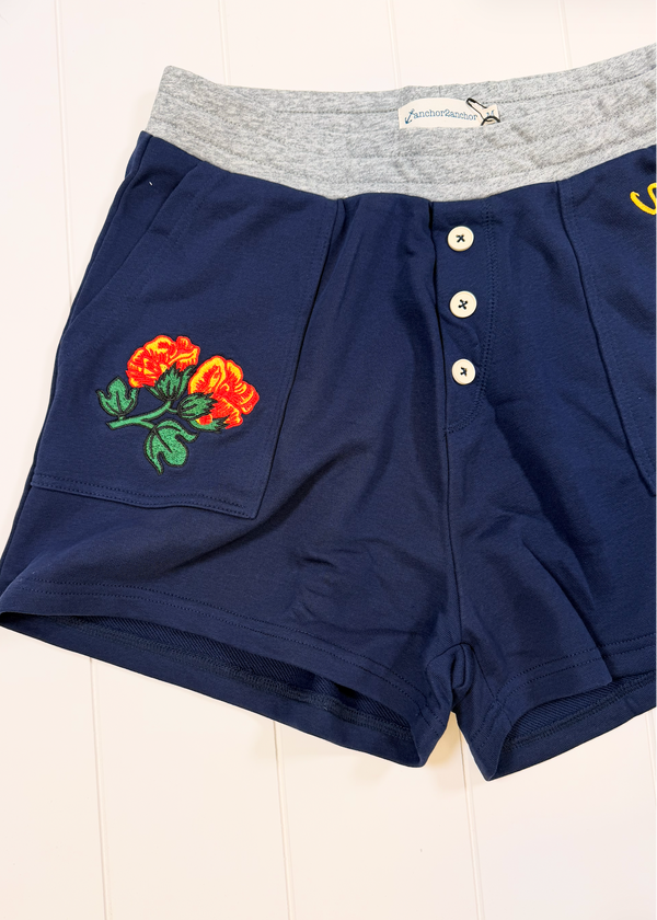 Navy blue shorts with a gray elastic waistband, featuring a three-button front closure, side pockets, and an embroidered orange rose on the left leg.