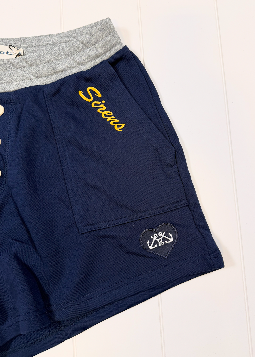 Navy blue shorts with a heather gray elastic waistband, featuring 'Screws' embroidered in yellow on the chest pocket and a black heart patch with a white anchor on the lower leg.