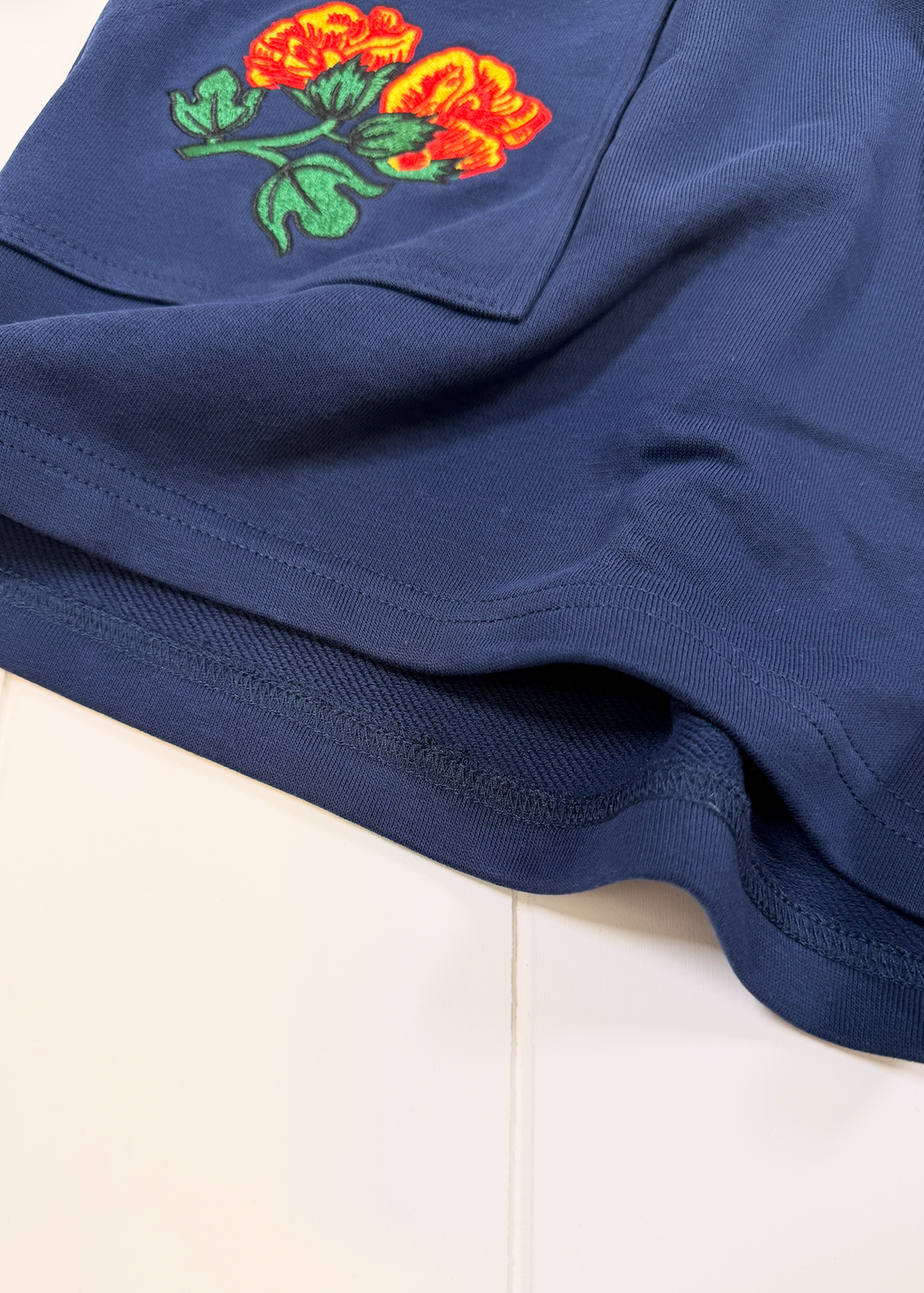 Close-up of a navy blue garment, likely shorts or a skirt, featuring an embroidered floral patch with orange-red roses and green leaves on the upper left side, laid flat on a white surface.