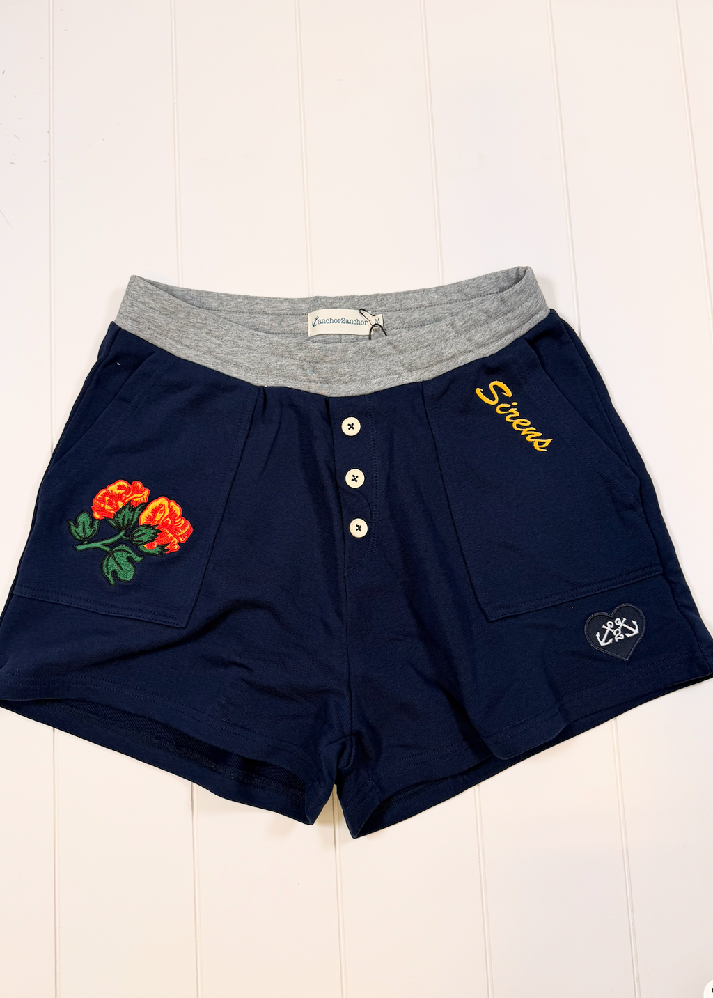 Navy blue shorts with a heather gray elastic waistband, featuring a three-button placket, embroidered orange roses on the left leg, yellow 'Stroops' text on the right leg, and a small heart patch on the lower right leg.