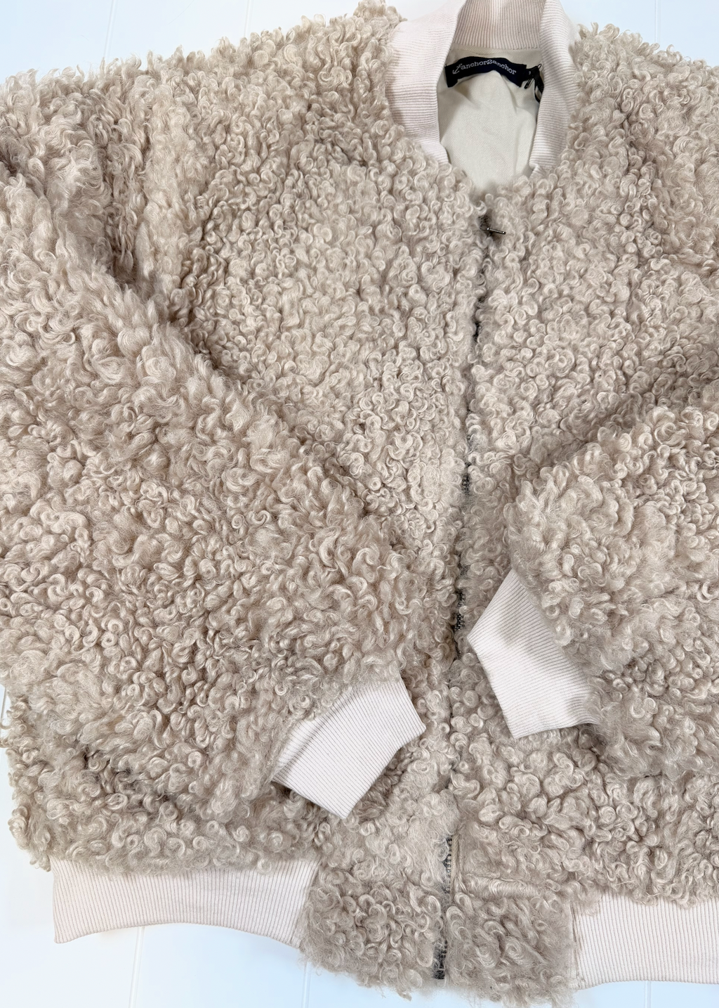 A beige curly sherpa fleece zip-up jacket with ribbed cream cuffs and collar, laid flat on a white background.