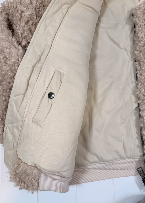 Close-up of a quilted beige jacket with a cream sherpa lining and a fluffy beige fleece collar, showing a side pocket with a snap button.