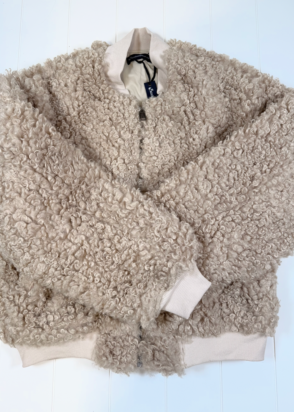 A beige sherpa fleece zip-up jacket with ribbed cream cuffs and hem, laid flat on a white wooden background.