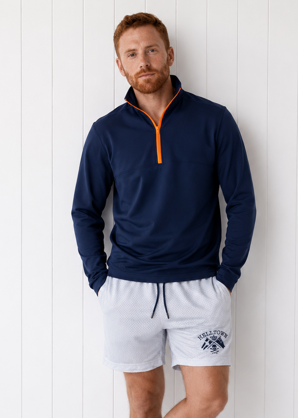 Man wearing a navy long-sleeve shirt with orange zipper and white shorts against a white wooden background