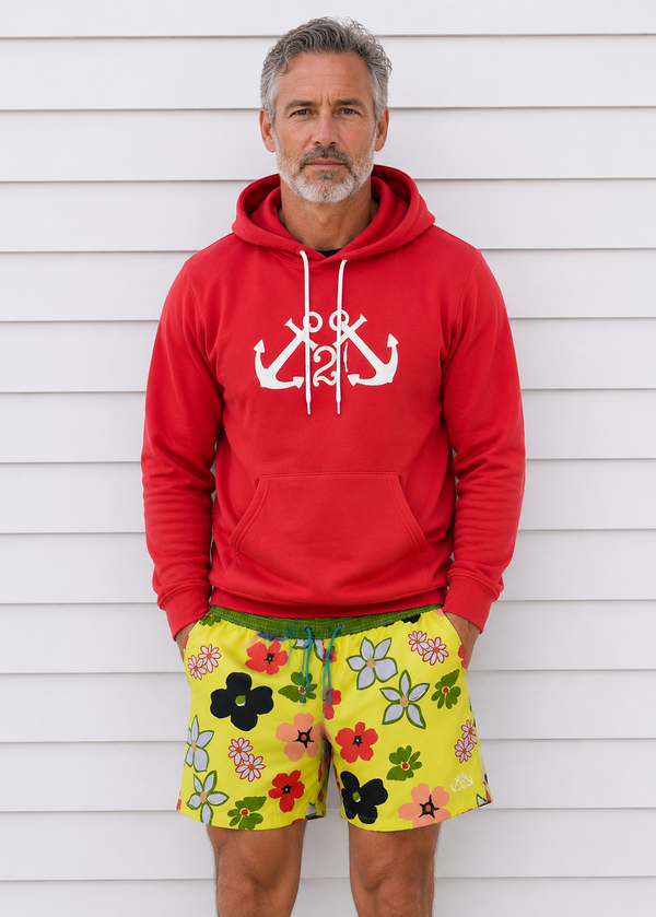 Man wearing a red hoodie with anchor design and colorful floral shorts against a white wooden background