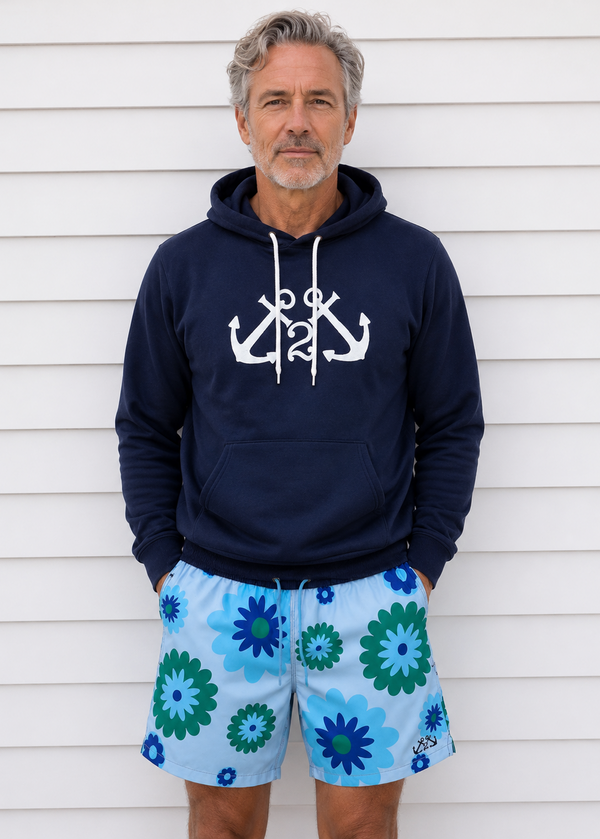 Man wearing a navy hoodie with anchor design and blue floral shorts against a white wall.